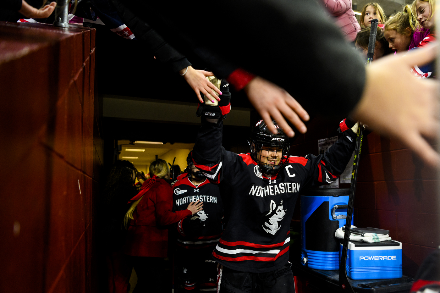 A hockey player in uniform and a helmet high fives fans leaning over the tunnel entrance as the player enters the arena