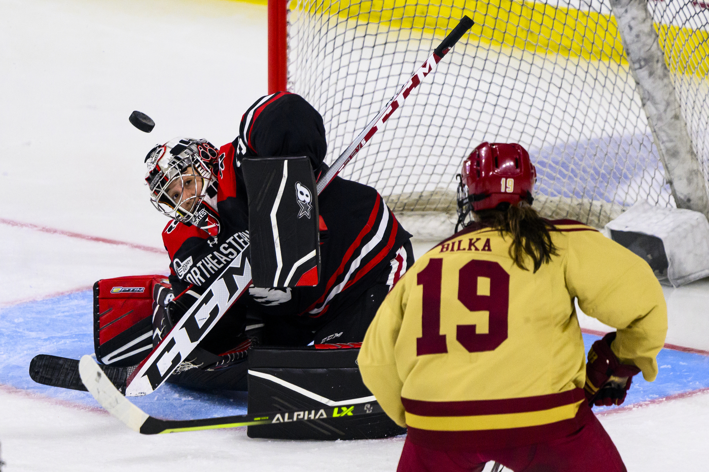 A Northeastern women's goalie looks up at a hockey puck that is flying towards her in the air as a BC hockey player looks on. 