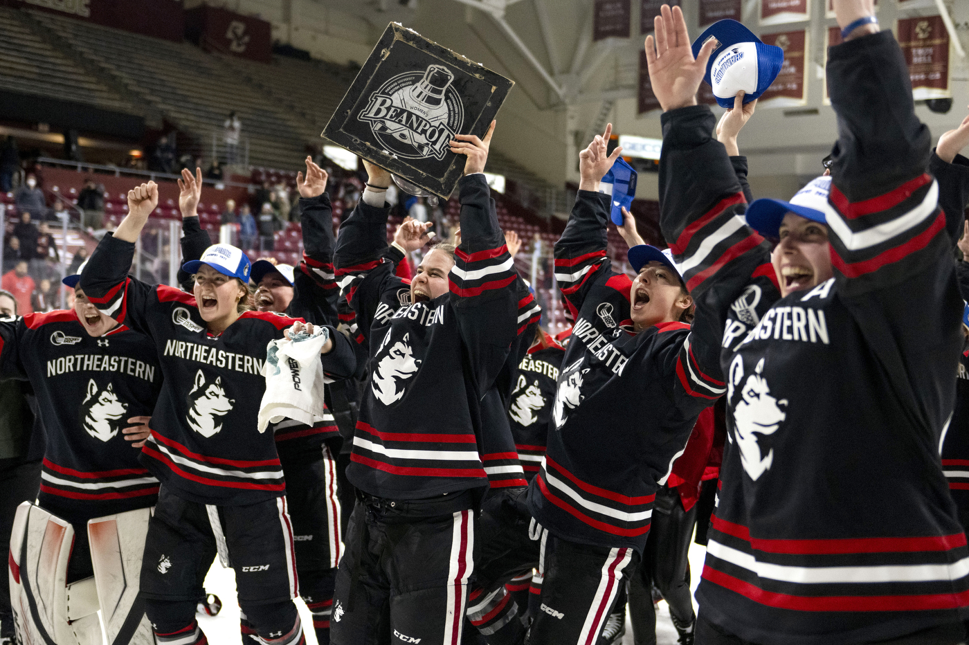 A line of players hold their hands up in celebration as one player holds the Beanpot trophy over her head