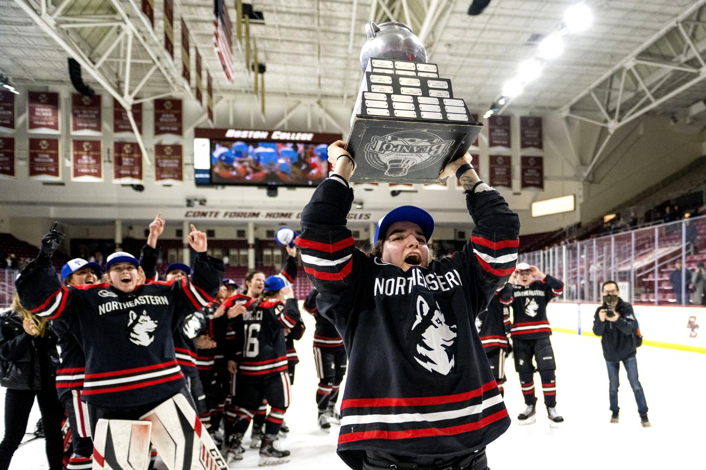A Northeastern women's hockey player hoists the Beanpot tropy over her head and smiles as her teammates look on and cheer.