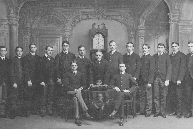 Skull and Bones Club of Yale photo, 1884, A group of men posing for a photo in front of a clock, with a skull on a table.