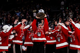 A Northeastern men's hockey player hoists the Beanpot trophy over his head and smiles as his teammates look on and cheer.