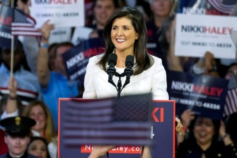 Nikki Haley stands behind a podium, smiling. A crowd looks on from behind wither several people holding up signs that say 'Nikki Haley'