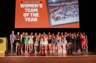 Members of the women's ice hockey team stand on stage in front of a screen that displays a photo of them on the ice and their award title.