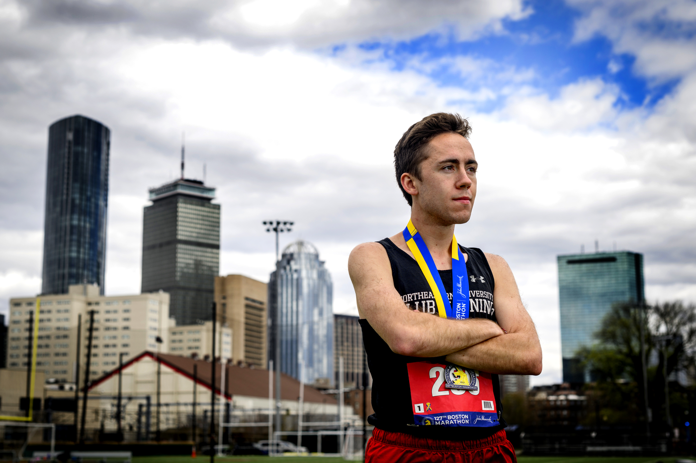 A runner with a medal around his neck stands with arms crossed in front of the Boston skyline.