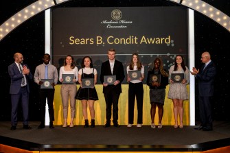 Students pose on a stage while holding awards