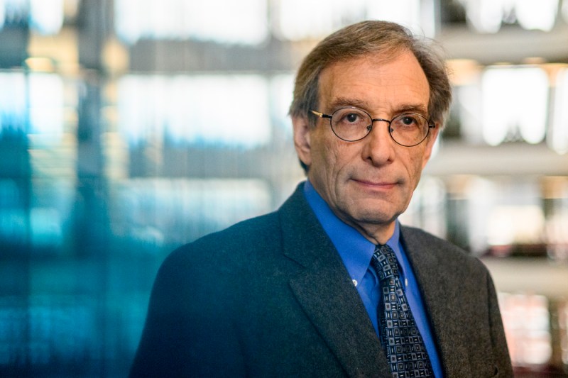 Professional portrait of a person wearing glasses, a dark suit jacket, blue shirt, and patterned tie, photographed in an office or academic setting with a softly blurred background.