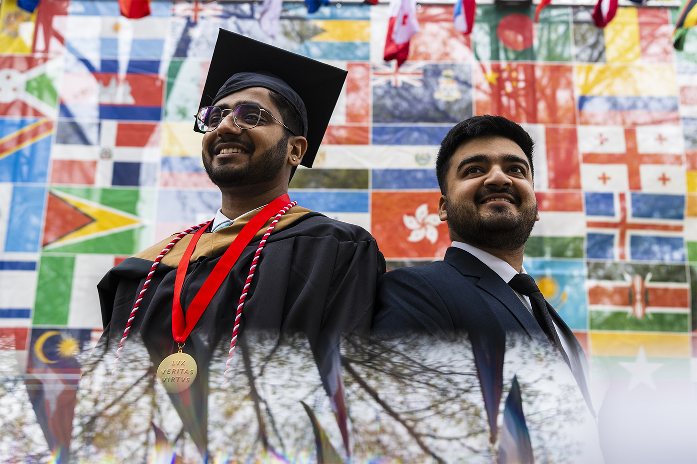 Two students, including one in cap and gown, pose against a backdrop of international flags
