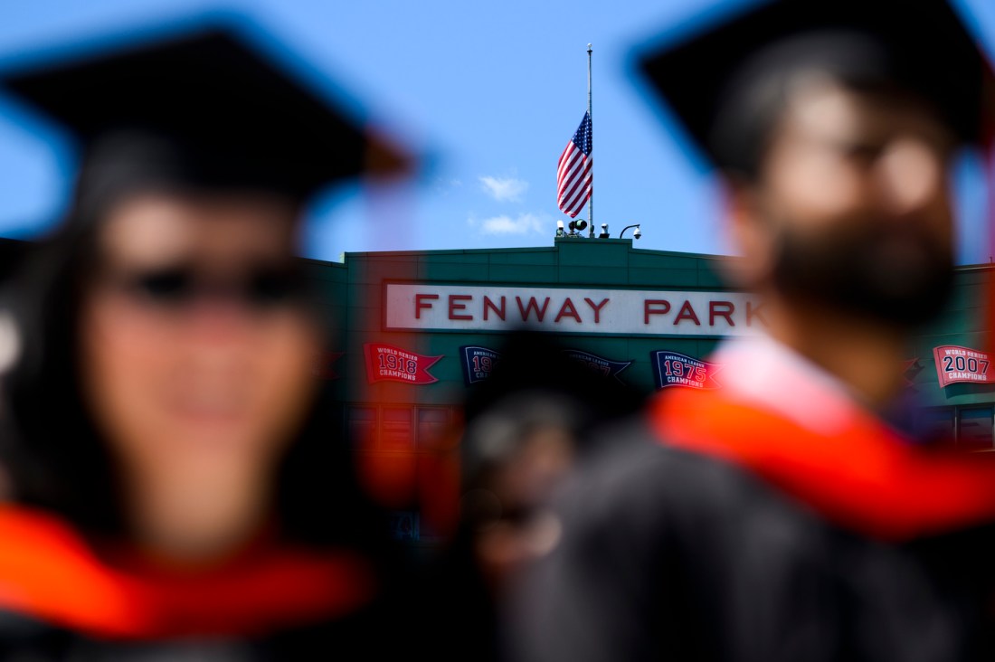 In the foreground, two graduates are blurry in their caps and gowns. In the background, the Fenway Park sign is on a wall of the park underneath a US flag.