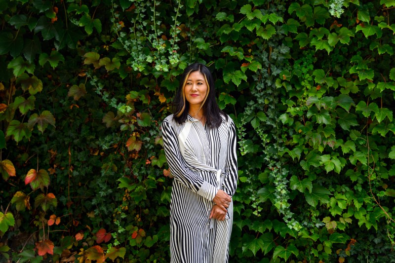 Denise Khor stands against a backdrop of lush green ivy in a black and white striped dress and with her arms crossed in front. 