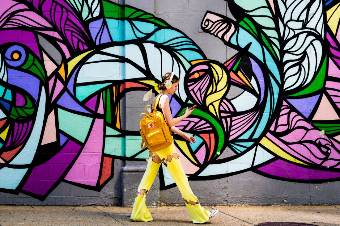 Person wearing yellow pants and headphones walking in front of a mural on campus.
