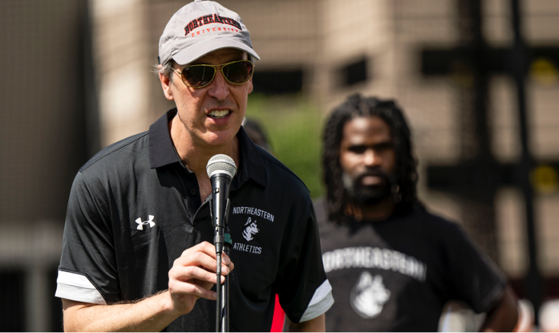 Michael Armini speaking at a microphone wearing a Northeastern Athletics shirt.