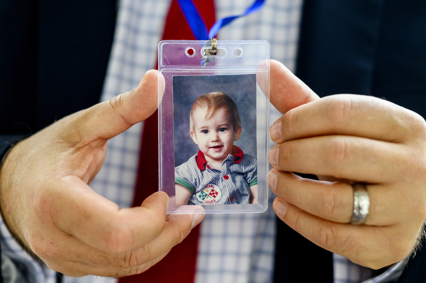 Detwiler, holding a lanyard to the camera--inside is a photo of his son, Riley. Riley looks to be a young toddler with red hair and dark eyes. He's wearing a striped shirt.