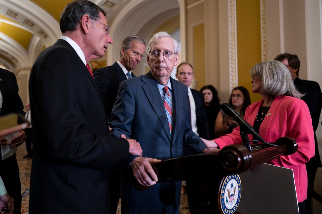 Mitch McConnell stands behind a podium at a news conference, while John Barrasso and Joni Ernst hold onto his wrists. Other officials stand behind Mitch McConnell.