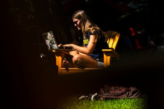 A person sits in an adirondack chair while working on a laptop. The background is blacked out.