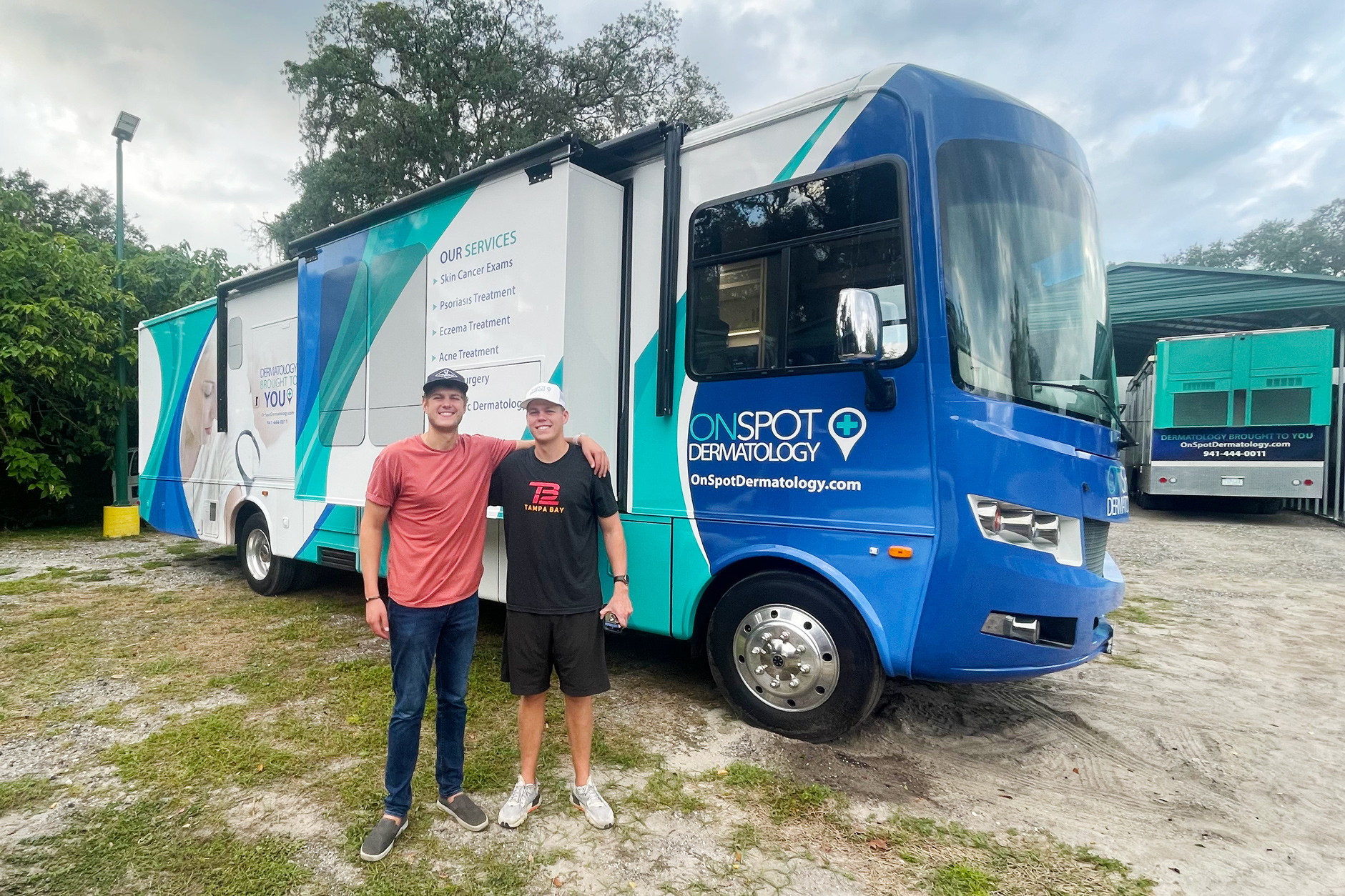 Brothers Darin, left, and Don Hunt, right,  pose for a photo in front of a blue and teal mobile dermatology clinic parked on a grassy lot in Florida.