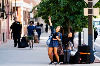A student takes a selfie and another person with several pieces of luggage on the curb near a tree.