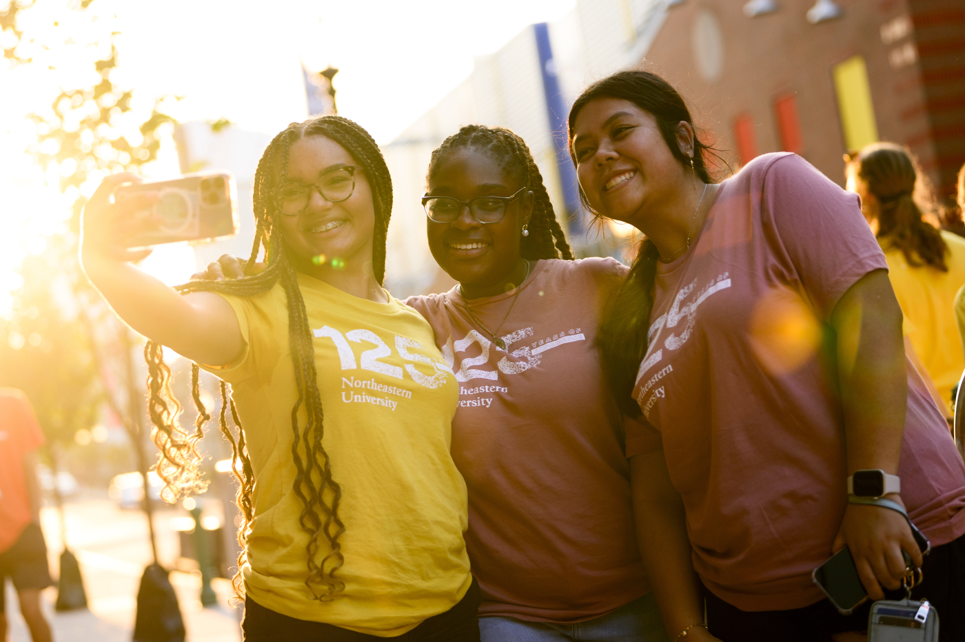Three students wearing t-shirts that say "125 Northeastern University" smile, while one student takes a selfie with her cellphone.