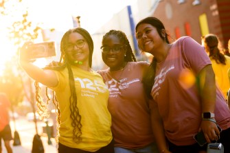 Three students wearing t-shirts that say "125 Northeastern University" smile, while one student takes a selfie with her cellphone.