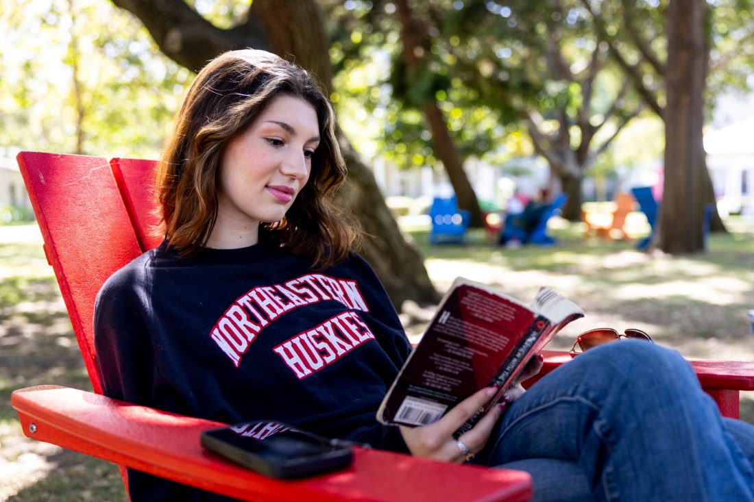 Person wearing a Northeastern huskies sweater reading a book in a red adirondack chair. 