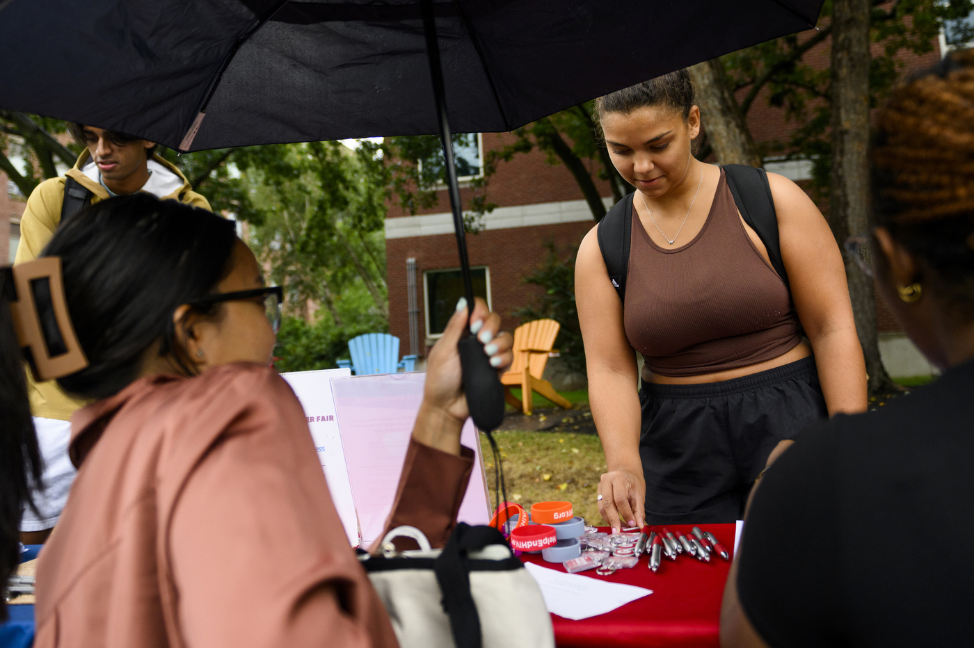 Student underneath a black umbrella approaches a table at the volunteer fair. 