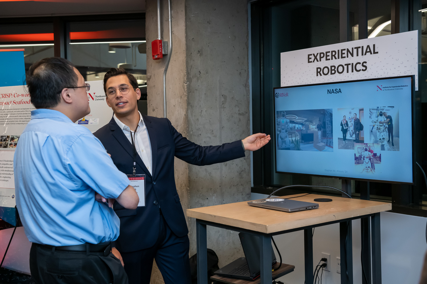 Speaker gesturing to a TV screen displaying a slide about Experiential Robotics at the Experience Powered by Northeastern campaign stop in Arlington.