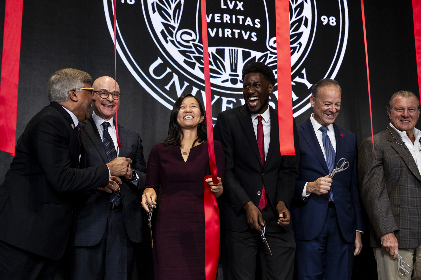 Six public figures laugh together while cutting red ribbons on a stage.