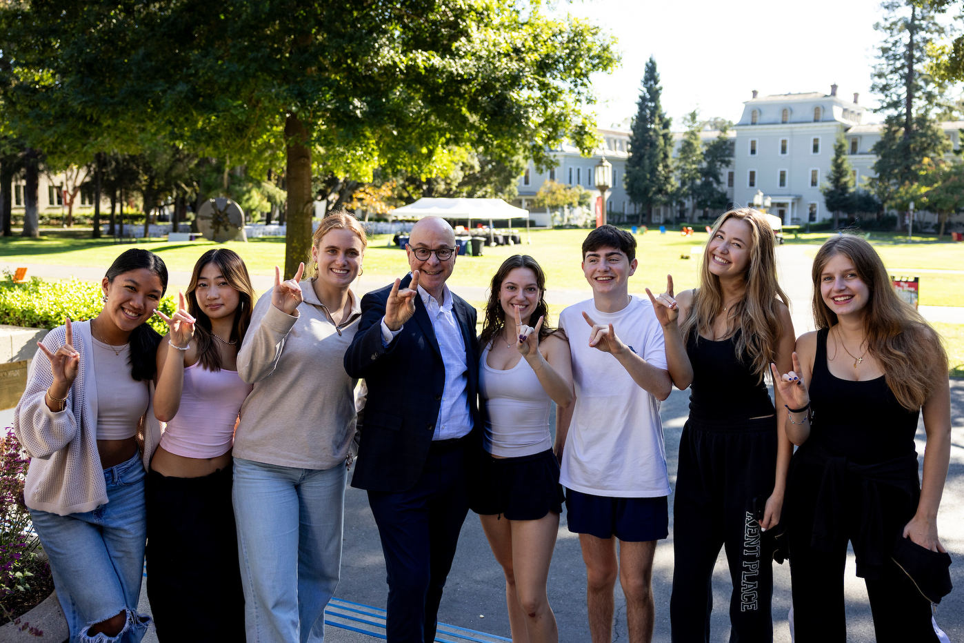 President Aoun posing with a group of students all doing a hand sign with their two middle fingers touching their thumb and their index finger and pinky raised. 