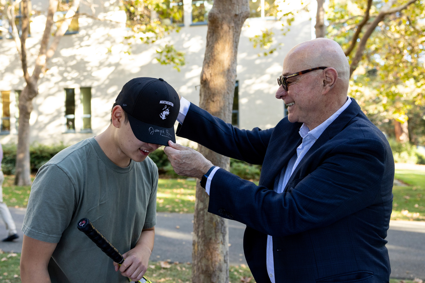 President Aoun putting a Northeastern branded baseball cap with his autograph on a student's head.