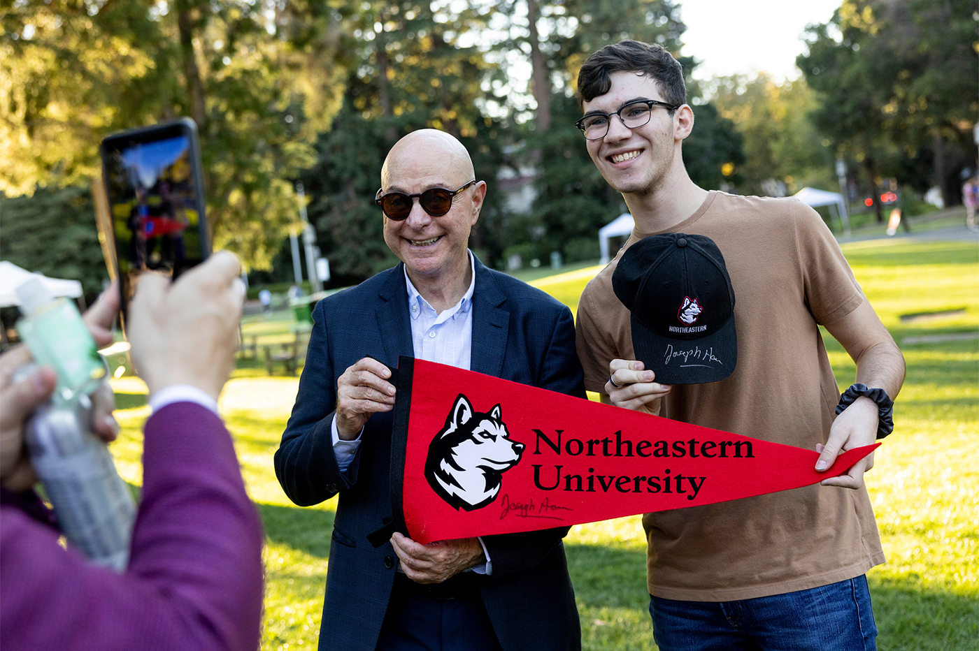 Student posing with a Northeastern branded, President Aoun autographed baseball cap next to President Aoun.