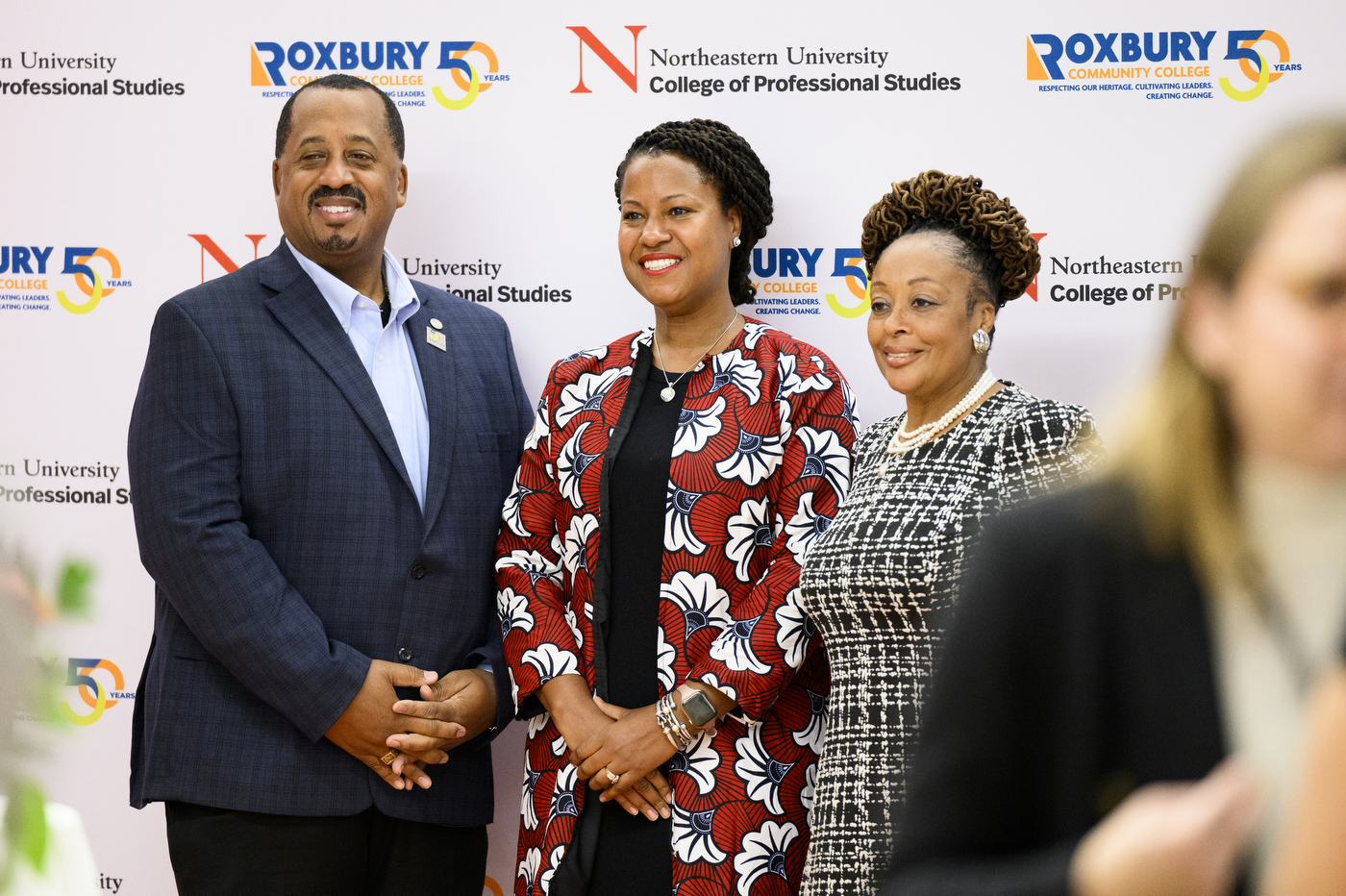 Three people posing together in front of a photo background with Northeastern University College of Professional Studies and Roxbury Community College logos on it.