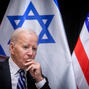 Biden pausing and looking pensive in front of both a US and Israel flag at a meeting.