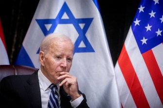 Biden pausing and looking pensive in front of both a US and Israel flag at a meeting.