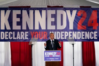 RFK Jr speaking during a campaign event with a banner behind him that reads 'KENNEDY24 DECLARE YOUR INDEPENDENCE' in all caps.