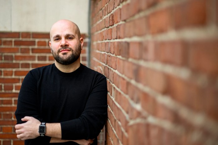 A bald man with a beard is leaning against a brick wall. He is wearing a black shirt.