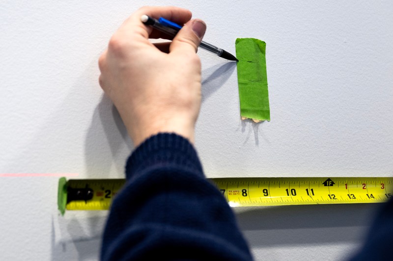 Person using green tape, a pencil, and a measuring tape to mark where art should be hung on the wall of the Gallery 360 exhibit. 
