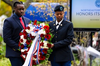 Two people, one in uniform, carry a wreathe of red, white, and blue flowers to honor lives lost.