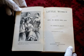Gloved hand holding open the title page of Louisa May Alcott's book Little Women.