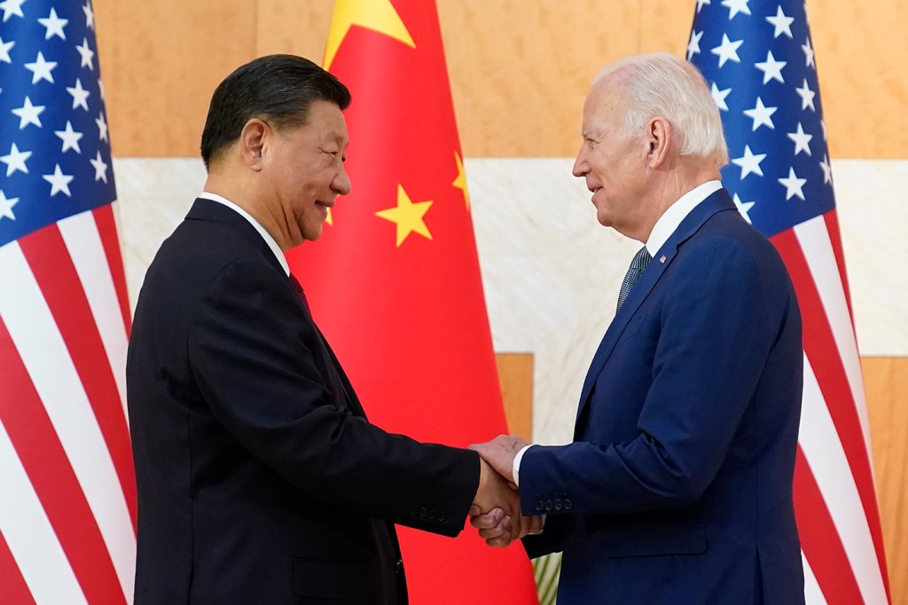 President Xi Jinping (left) and President Joe Biden (right) shaking hands in front of US and Chinese flags.