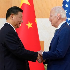 President Xi Jinping (left) and President Joe Biden (right) shaking hands in front of US and Chinese flags.