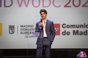 Isaac Moloney wearing a suit and speaking into a microphone at the World Universities Debating Championship.