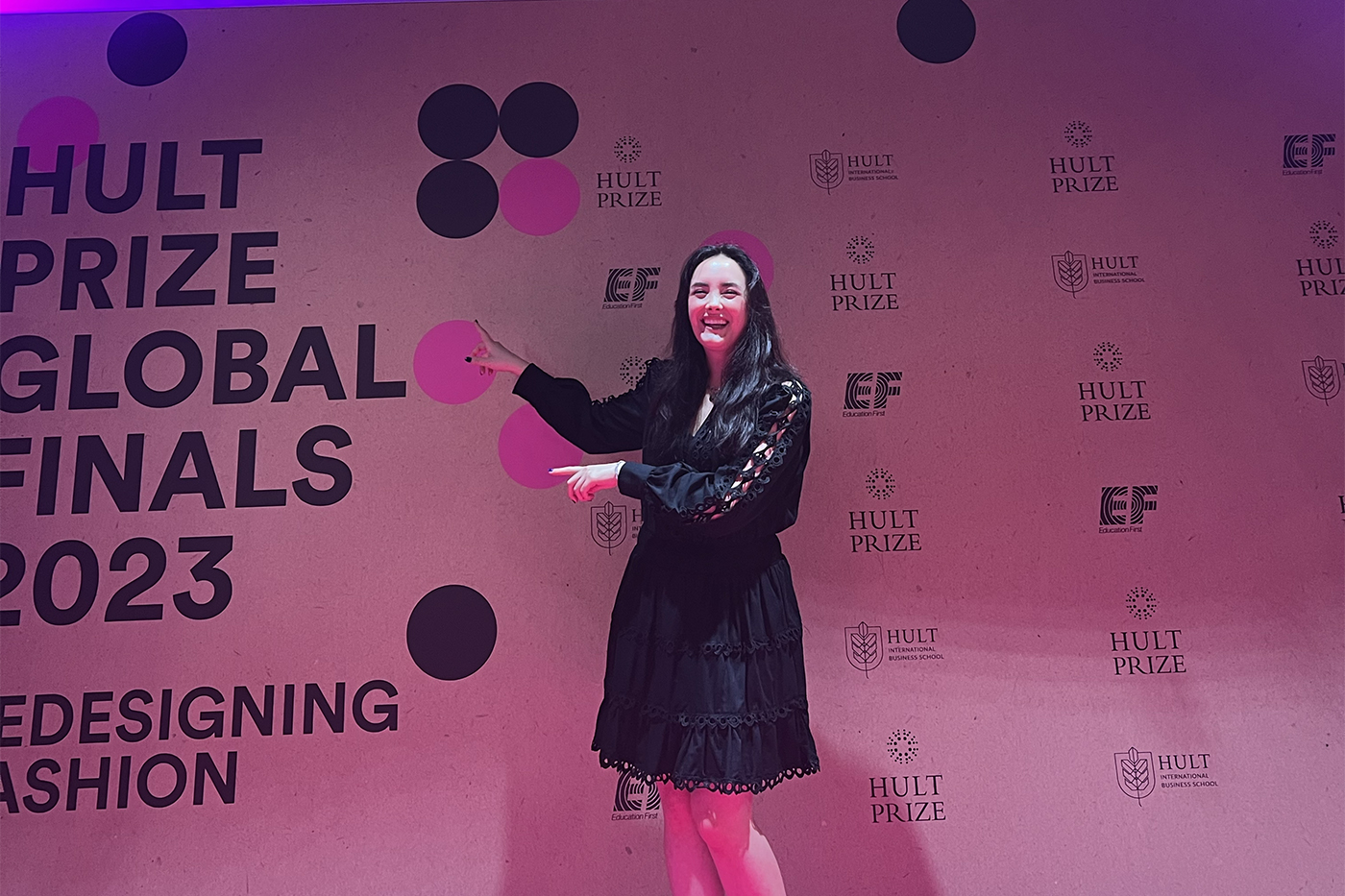 Izabella Pivo pointing to a photo background that says "Hult Prize Global Finals 2023, Redesigning Fashion".