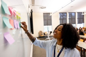 A person places multi-colored sticky notes on a white board inside a brightly lit classroom.