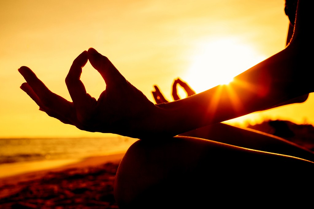 Silhouette of a person at sunset sitting cross legged with their hands resting in their lap, thumb and index fingers pressed together and palms upwards in Gyan mudra.