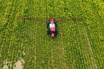 A red tractor is seen from above, spreading insecticides over a green field.