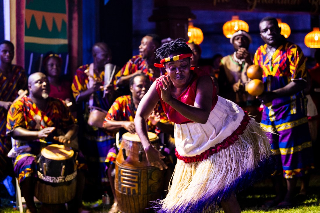 Ghana woman in traditional Ghana clothing dancing while other people play drums and make music behind her. 