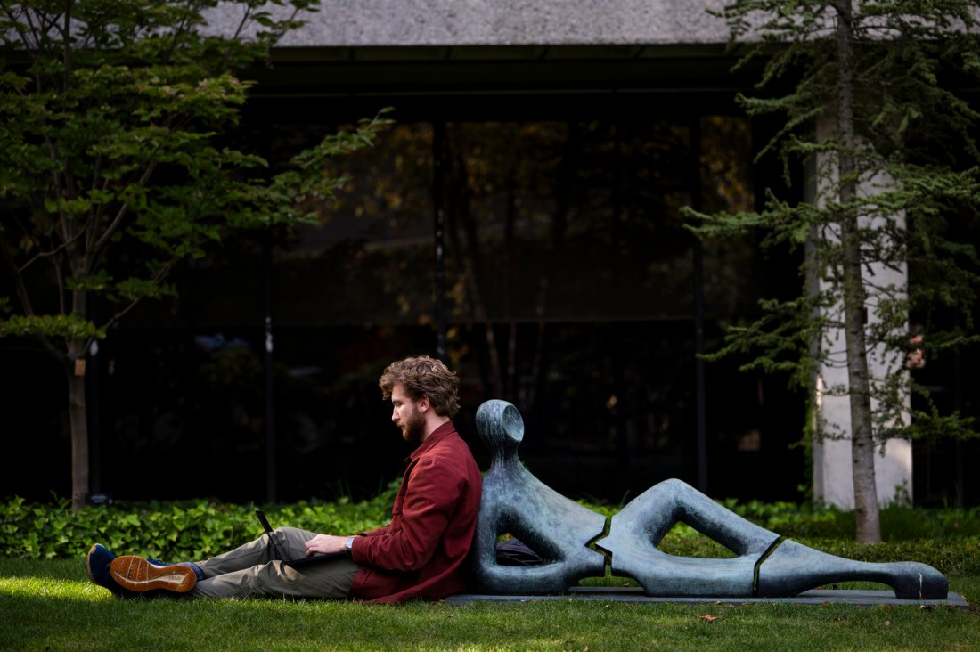 Theo Amendola leaning up against a sculpture while using his laptop. 