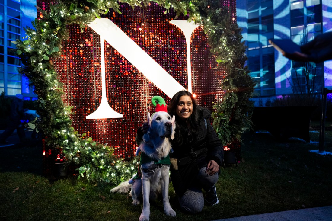 A person takes a picture with a dog wearing an elf-themed hat in front of a large white letter 'N' over a red background.