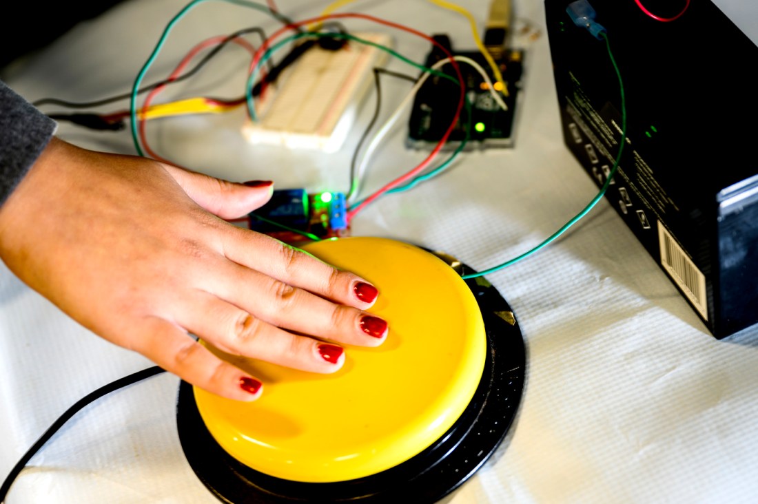 A student's hand with red nail polish pushes a large, yellow button, hooked up to various boards and small machines.