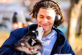 A person wearing headphones gets kissed by a husky puppy while outside.
